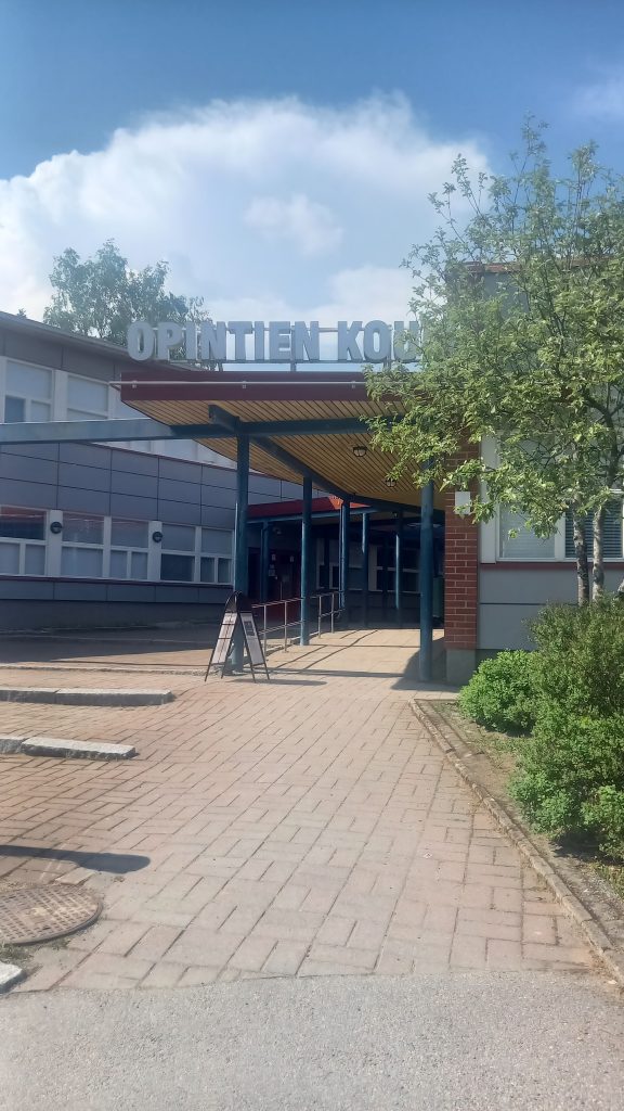 Kesäinen kuva Kärkölän Opintien koulun sisäänkäynnistä, josa näkyy rakennuksen seinä ja katos
